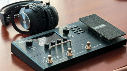 Una pedalera multiefectos digital con pantalla y un pedal de expresión, junto a unos auriculares sobre una mesa de madera.